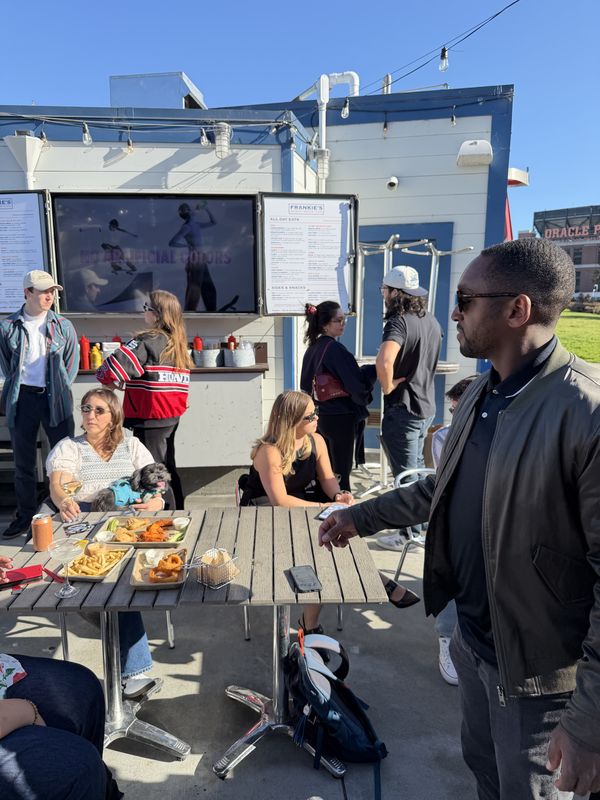 Attendees gathered around a patio table with shared plates of food on a sunny afternoon, with the ballpark in the distance.