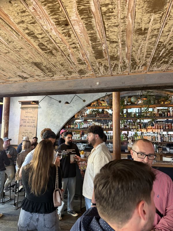 A packed bar under a hammered gold ceiling, with backlit shelves of bottles and guests chatting with drinks in hand.
