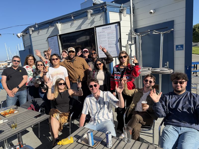 A big group of smiling professionals waving at the camera on a sunny waterfront patio, with sailboat masts in the background.